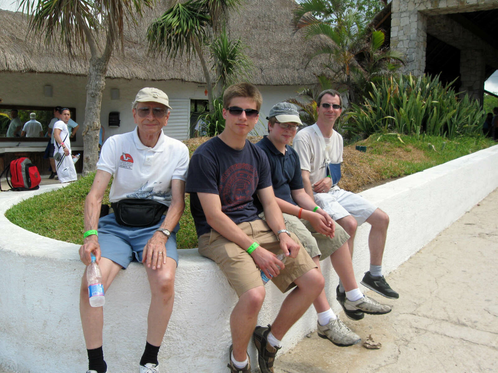 Four men sit on a white curved wall beside a tropical resort, posing for a photo with palm trees in the background.