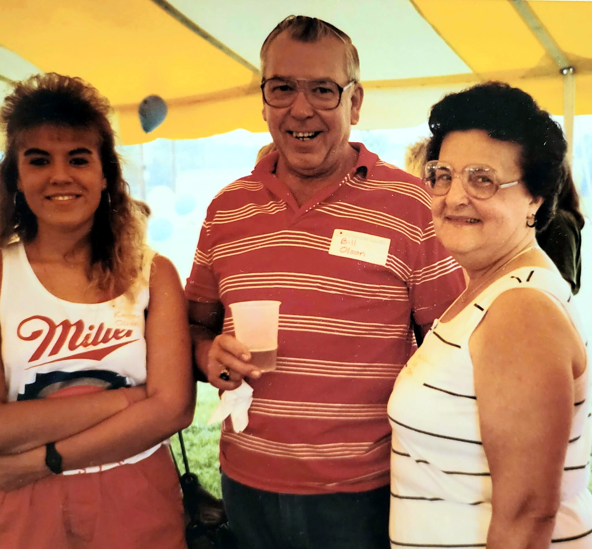 Three adults posing together at an outdoor event: a man in a red striped shirt with a name badge holds a cup between two smiling women.