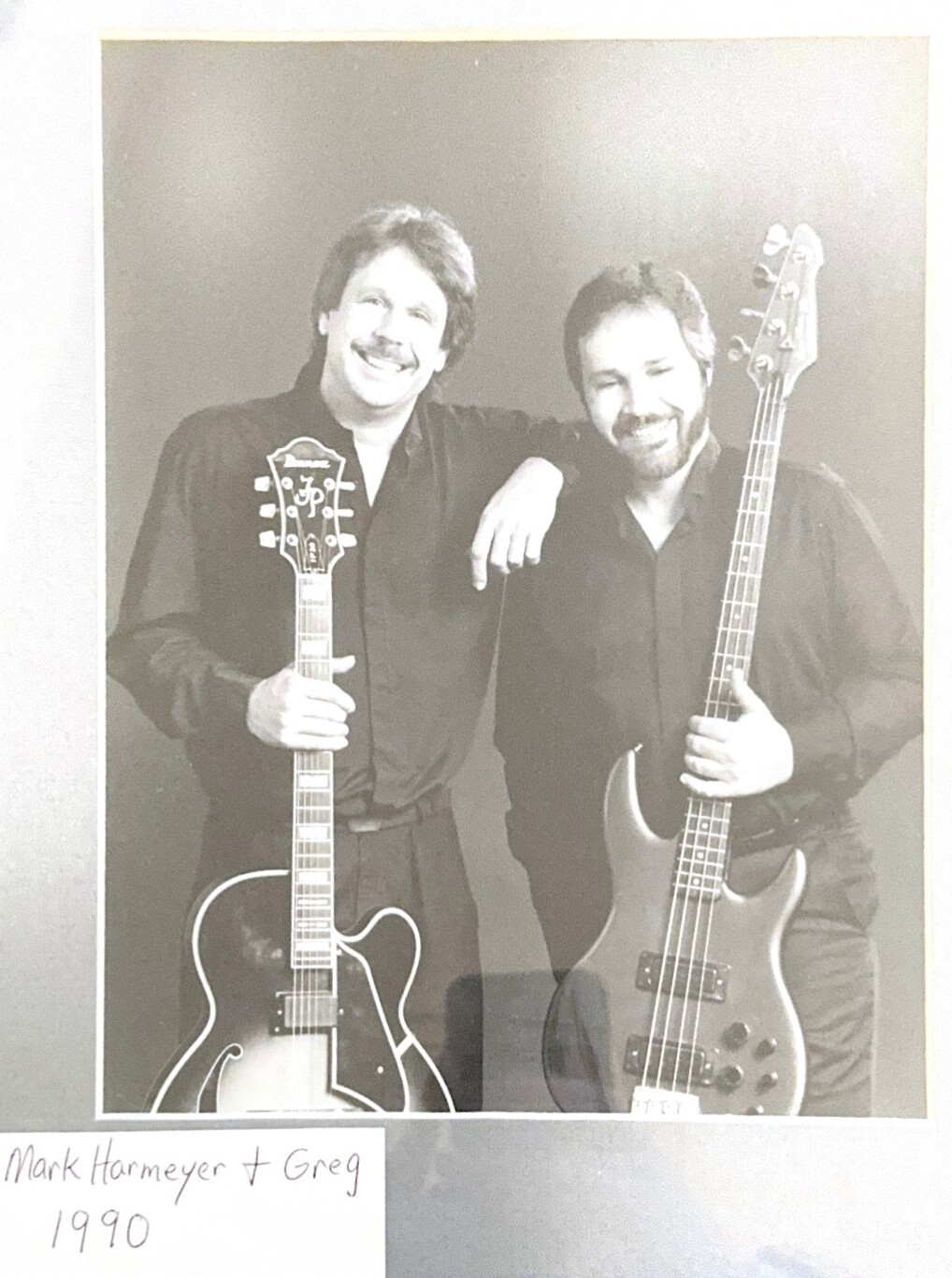 Two men in dark shirts pose with guitars, smiling at the camera; left man holds a hollow‑body guitar, right man holds a bass guitar, in a vintage black‑and‑white photo labeled 1990.