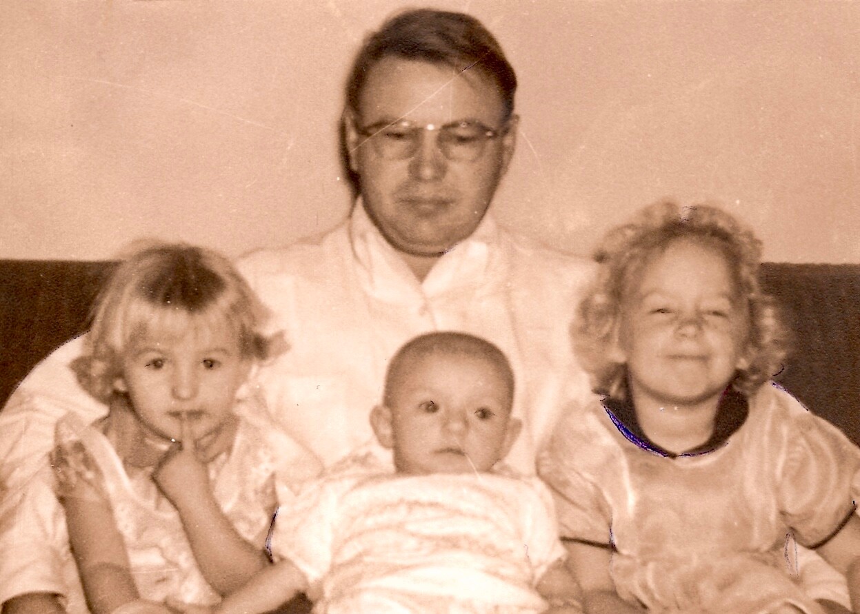 Man wearing glasses sits on a sofa with two young girls and a baby in front, in a sepia-toned family photo.