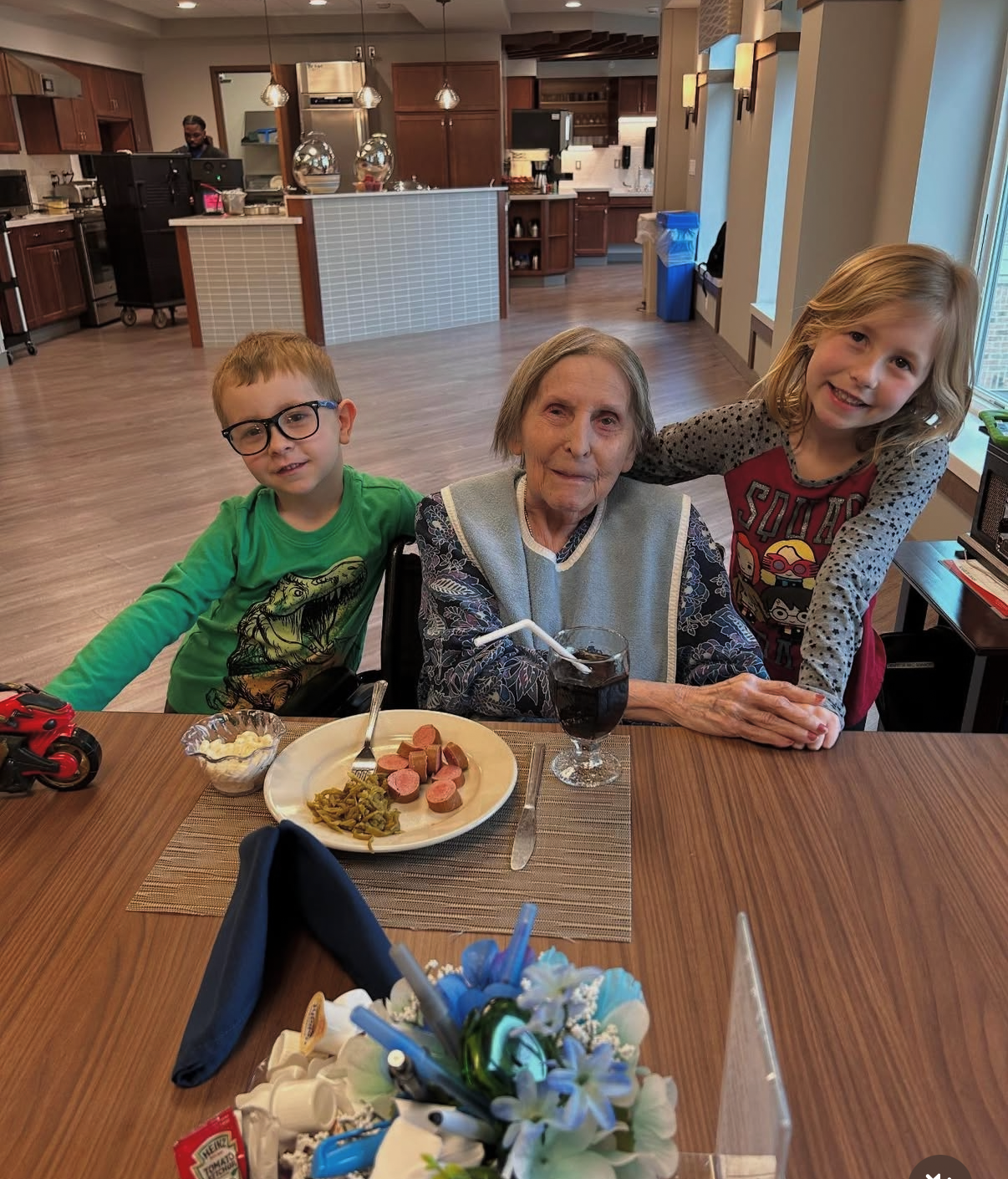 An elderly woman seated at a dining table with a young boy in a green shirt and a smiling girl leaning in from the right; food plate and drink in front, warm restaurant setting in background.