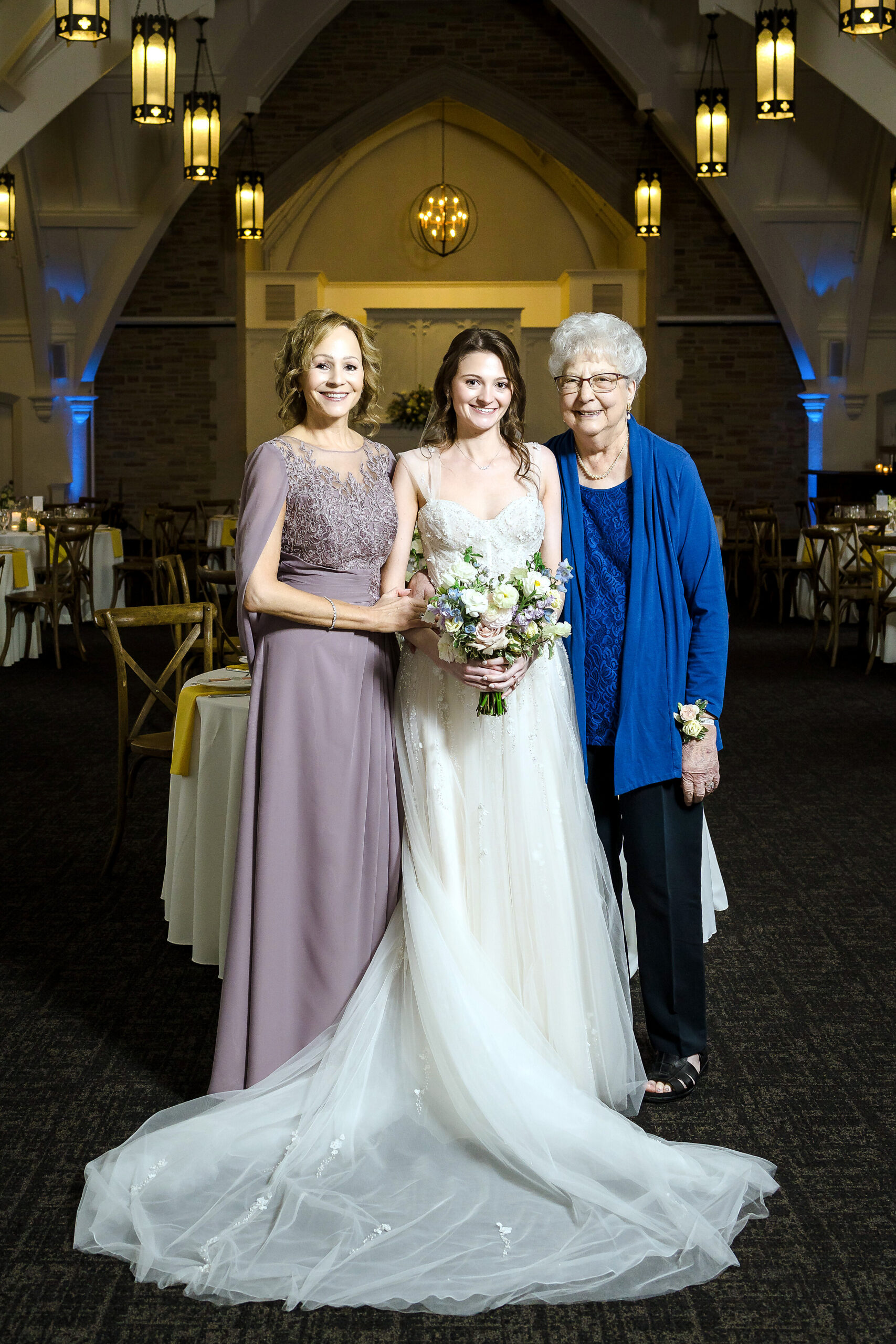 Bride in a white wedding gown holding a bouquet, flanked by two women in formal dresses inside a decorated venue with chandeliers.