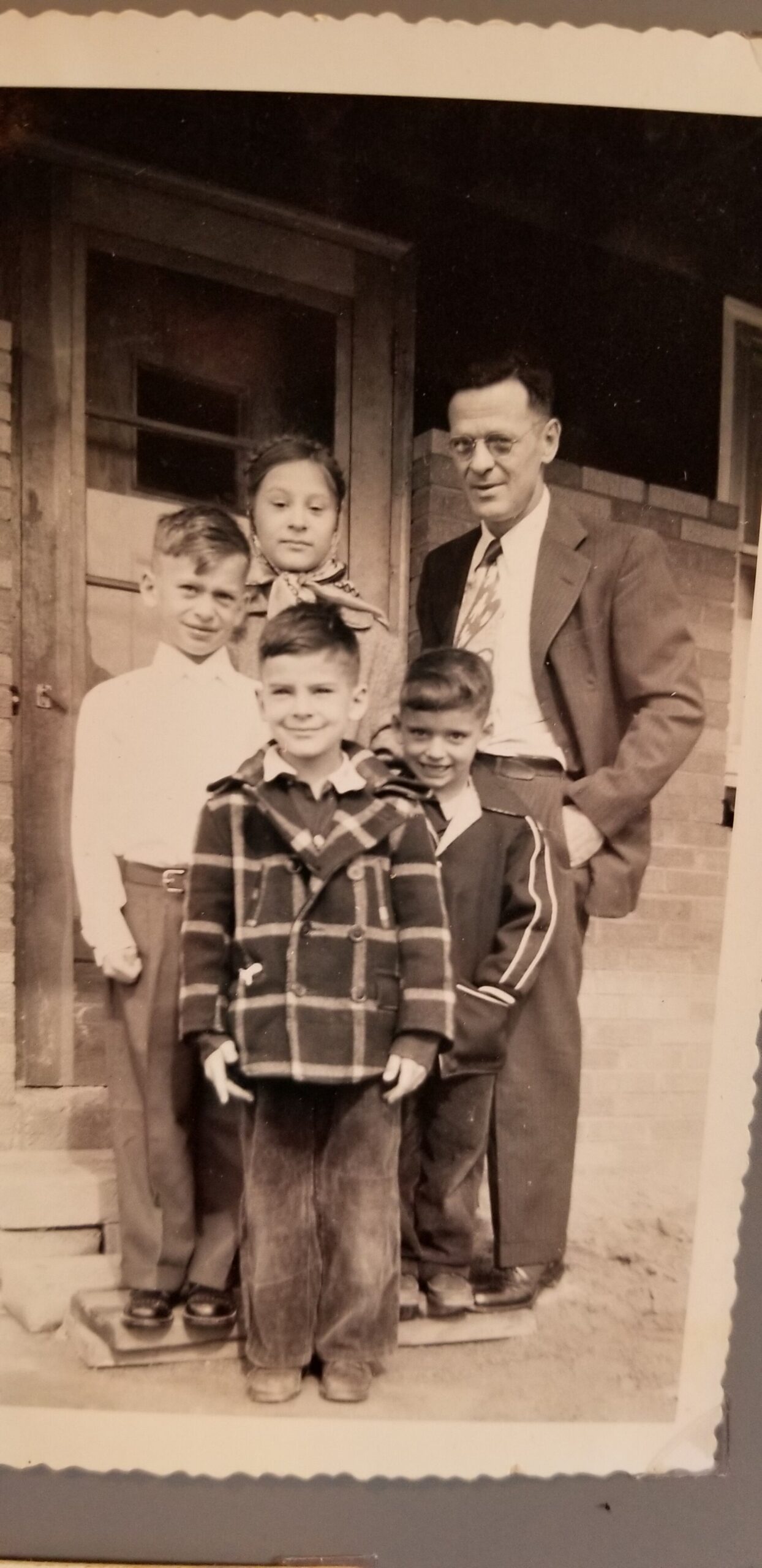 A man in a suit and glasses stands with four children on the front steps of a brick house, posing for a family photo.