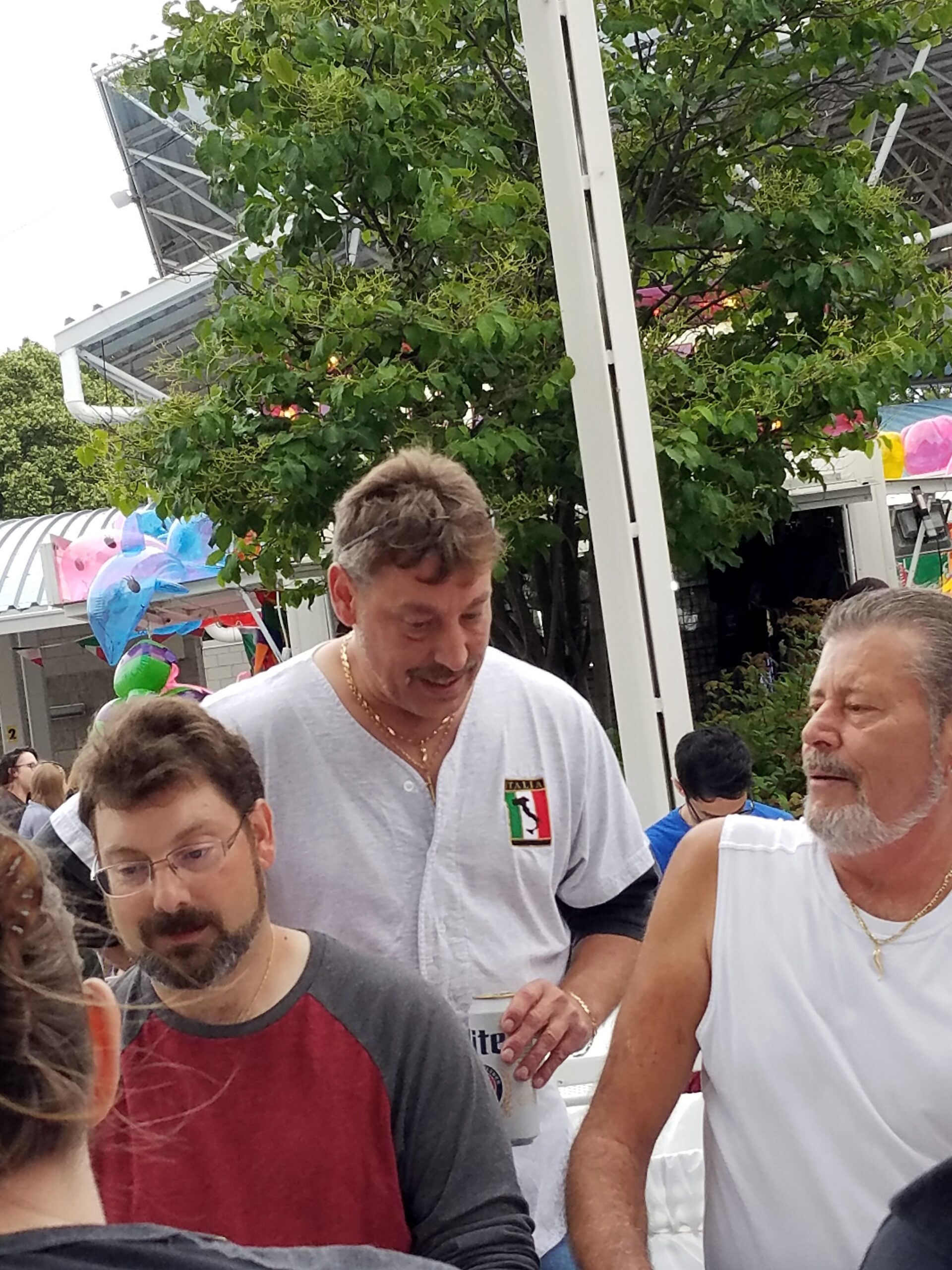 Group of people gathered outdoors, a man in a white shirt with an Italian flag badge speaks to others amid colorful balloons.