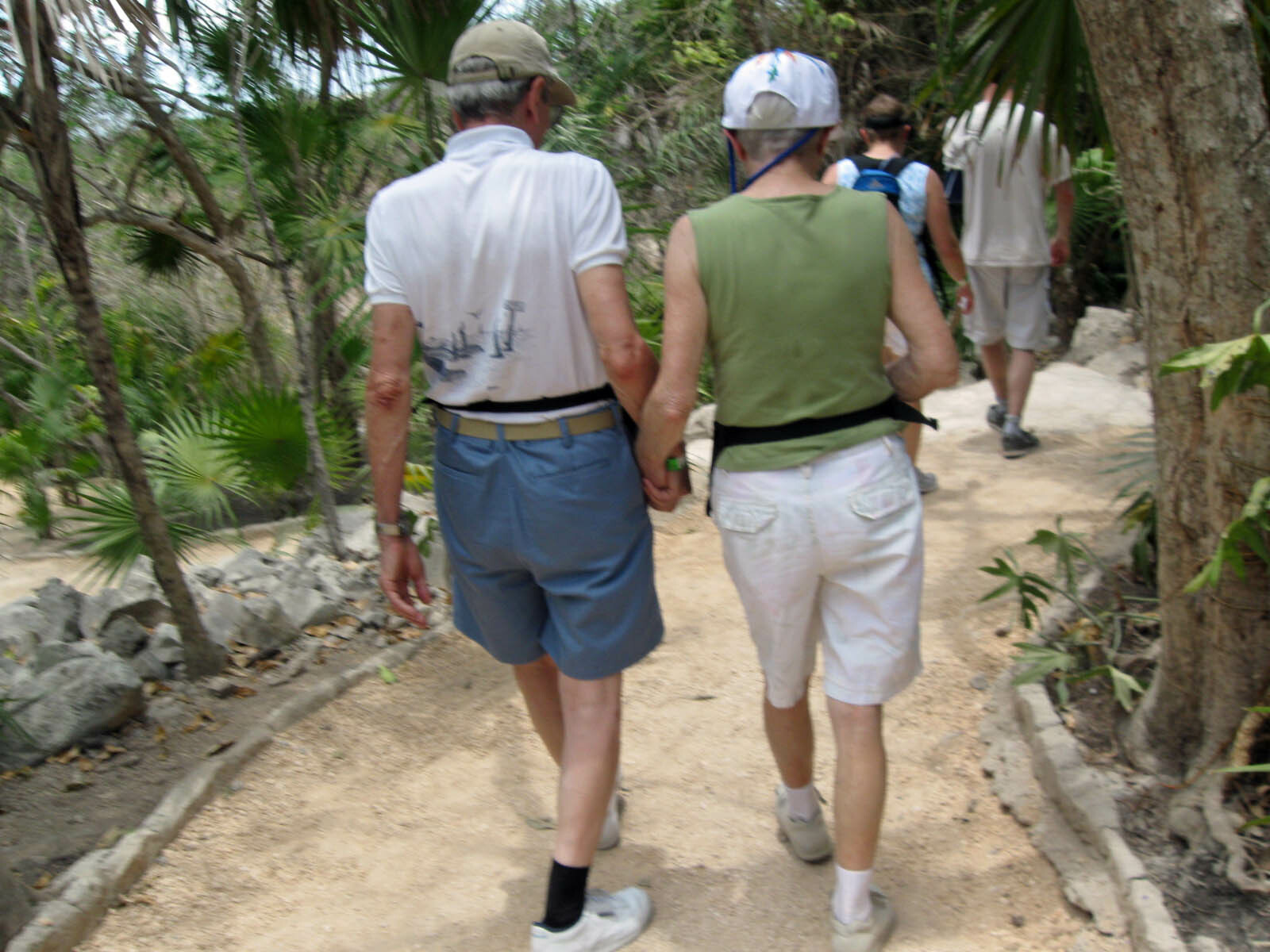 Two older hikers walk side by side on a dirt trail through a tropical, leafy area.