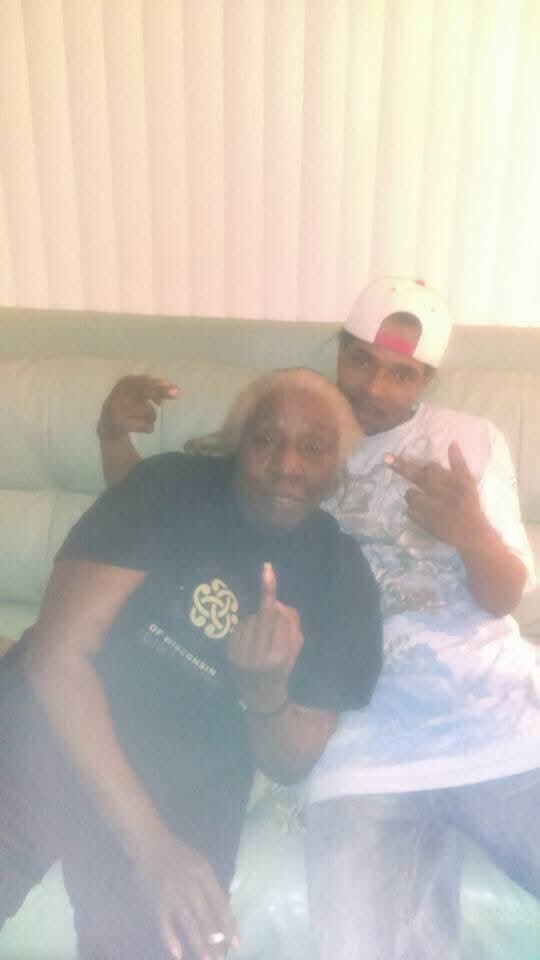 Two men sit on a beige couch, flashing middle fingers at the camera in a casual living room setting.