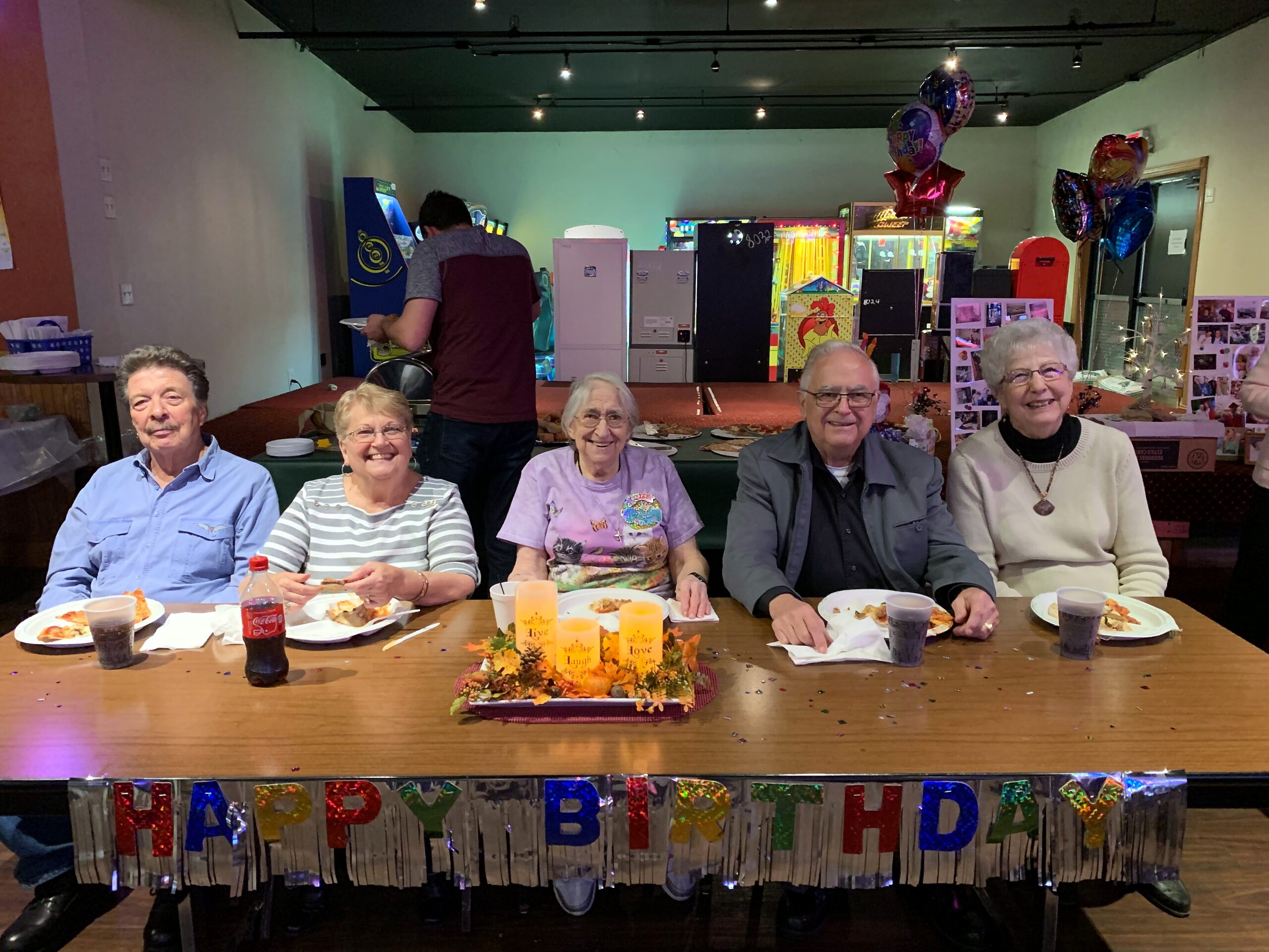 Group of five elderly people seated at a long table, sharing cake and drinks at a birthday party, with balloons and arcade machines in the background.