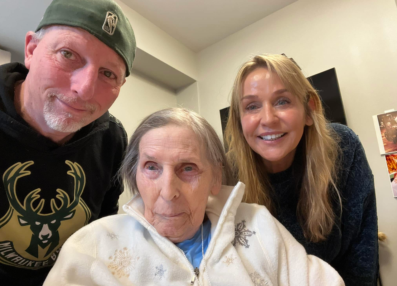 Three people pose for a selfie: an elderly woman in a white zip‑up sweater between a man in a green cap and a smiling blonde woman beside her in a home setting.