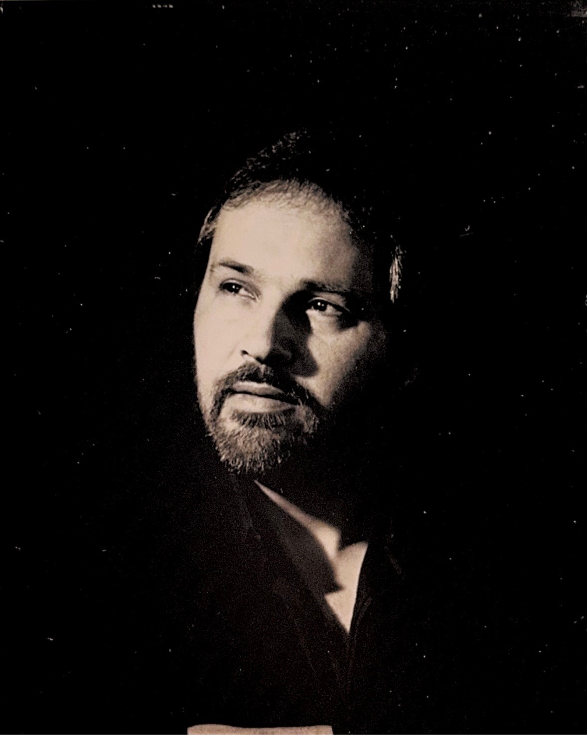 Portrait of a man with short dark hair and a trimmed beard, gazing to the right against a dark, speckled background.