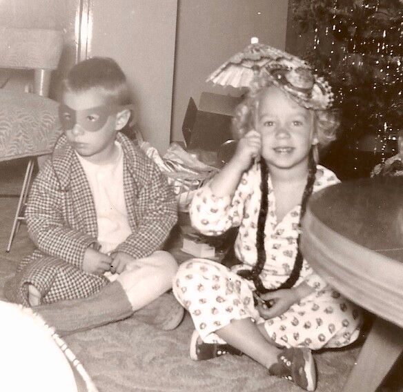 Two young children sit on the floor at a Christmas gathering: a boy in a robe and eye mask, and a girl in pajamas with a hat and braids, with a Christmas tree in the background.