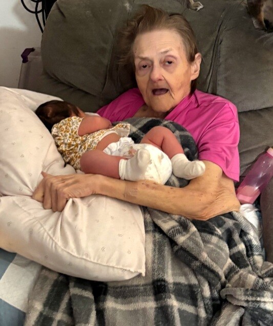 Elderly woman in a pink shirt sits up in bed, cradling a newborn baby wrapped in a blanket.