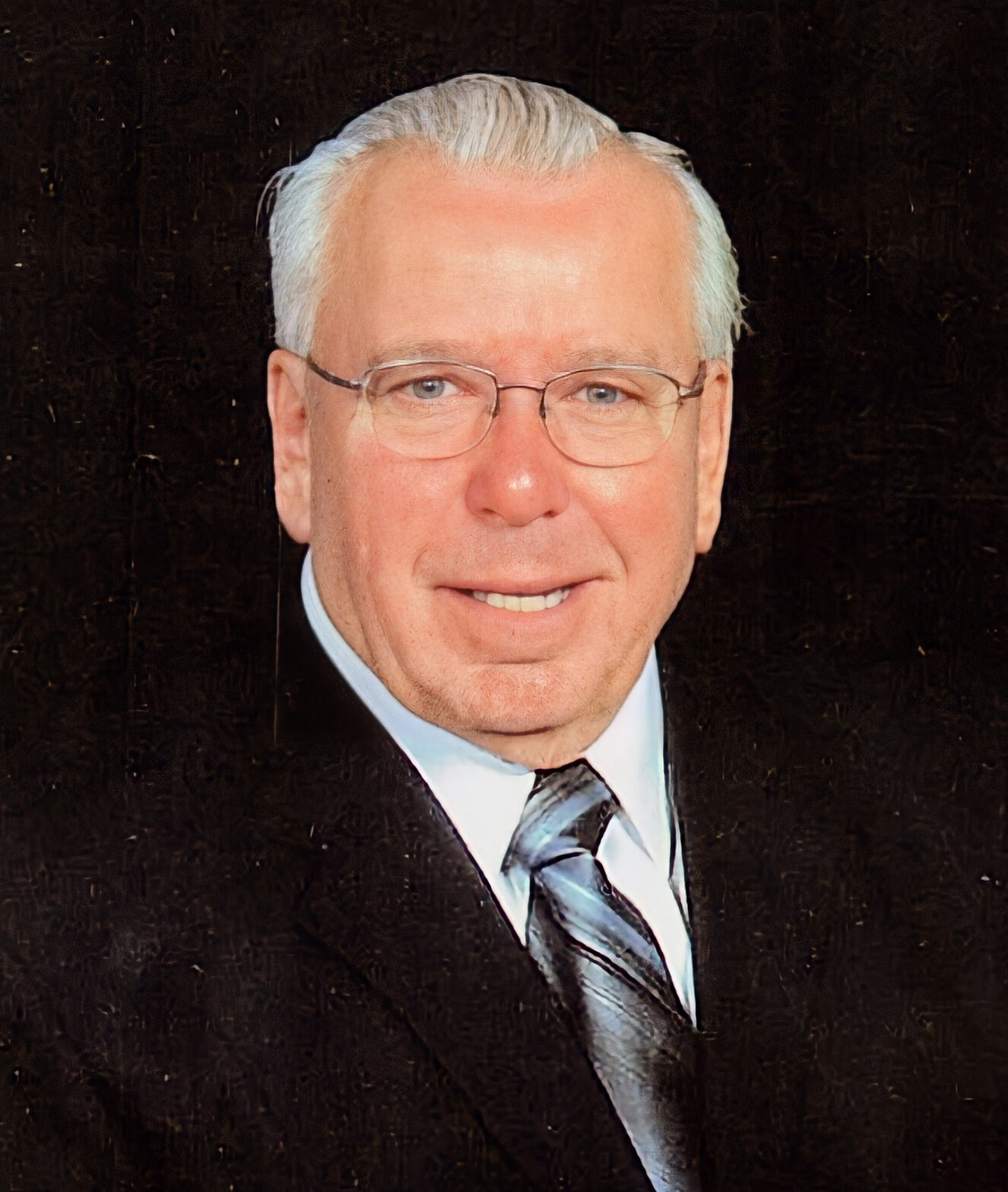 Smiling older man in a dark suit and striped tie, wearing glasses, posing for a formal portrait.