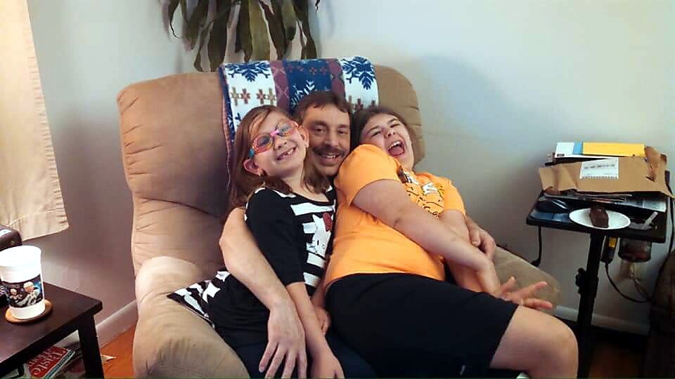 A man sits on a beige recliner with two smiling children leaning in for a hug on either side of him in a cozy living room scene.