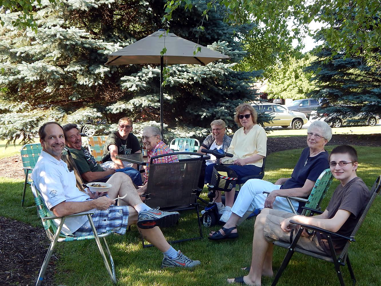 Group of eight adults and a teenager seated in lawn chairs around a table under a patio umbrella in a sunny backyard, smiling and talking.