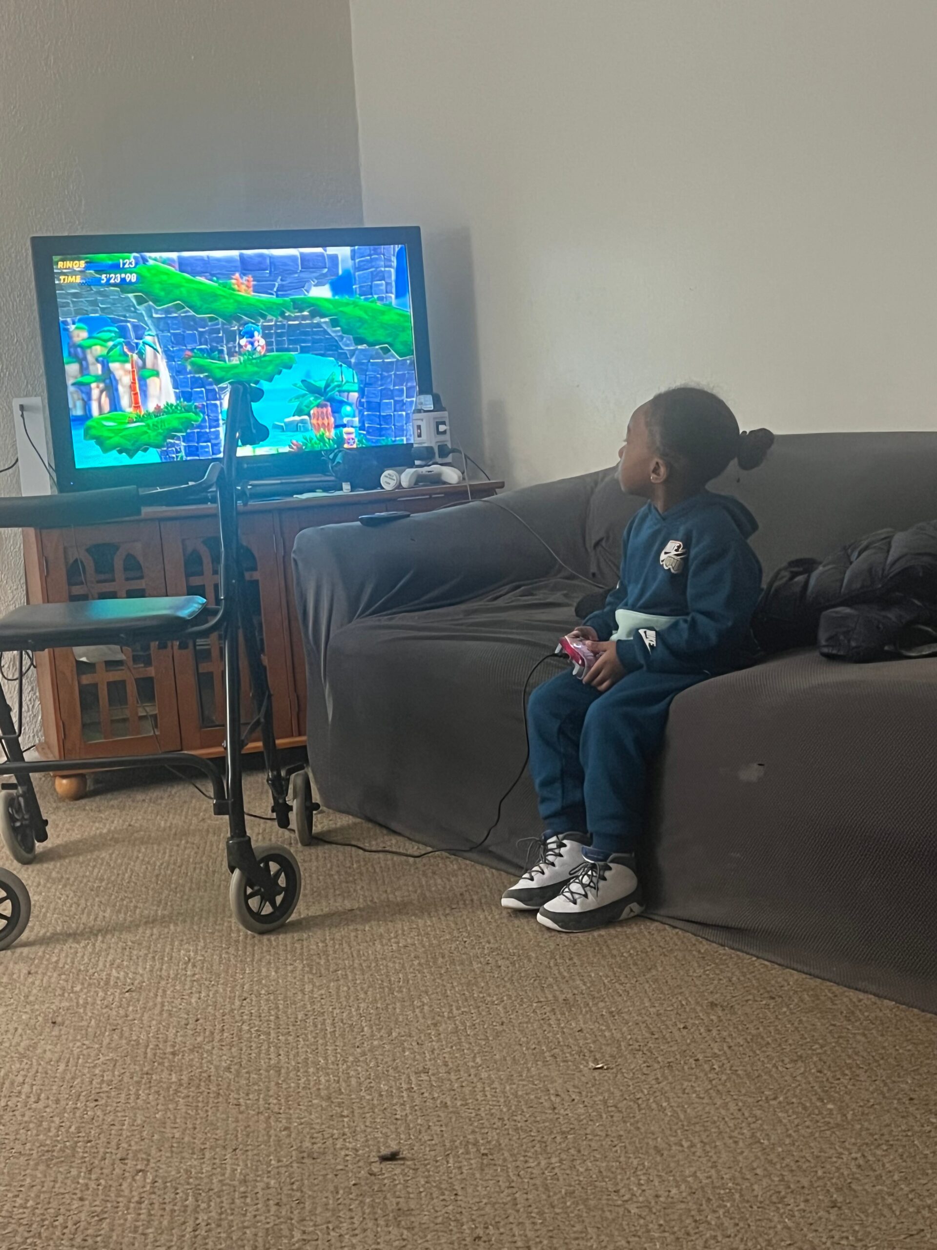 Child sits on a gray couch, holding a game controller and playing a bright platform video game on a large TV screen.