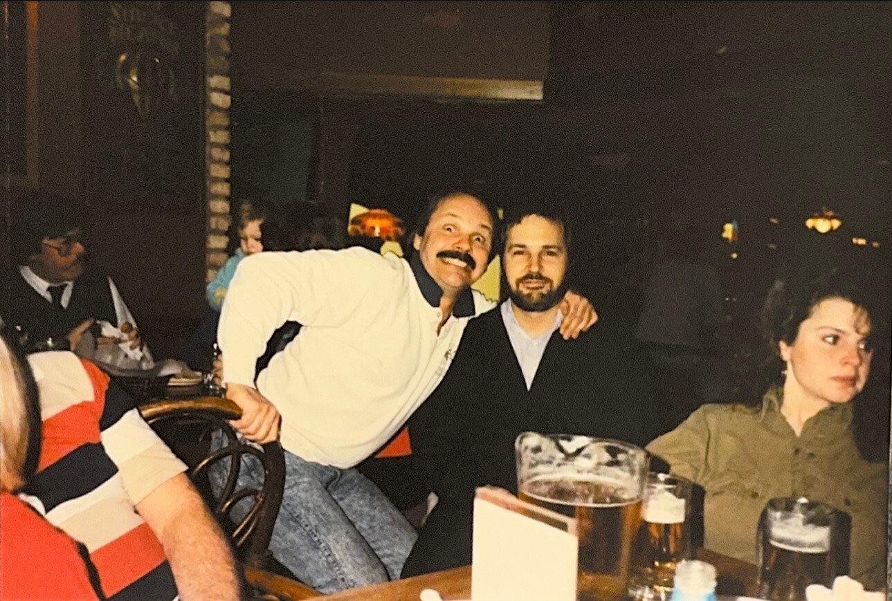 Two men posing with arms around each other at a restaurant table, with beers in front and a woman seated nearby looking away.
