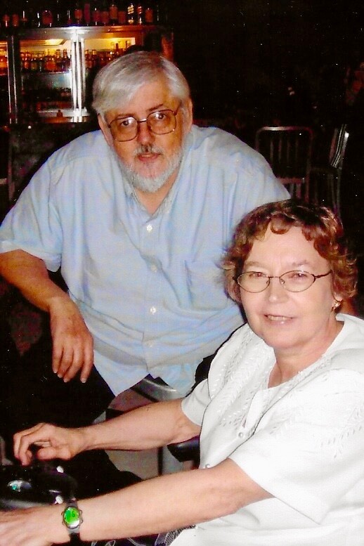 Older couple at a bar; man with white hair and beard in a light blue shirt, woman with glasses in a white blouse smiling at the camera.