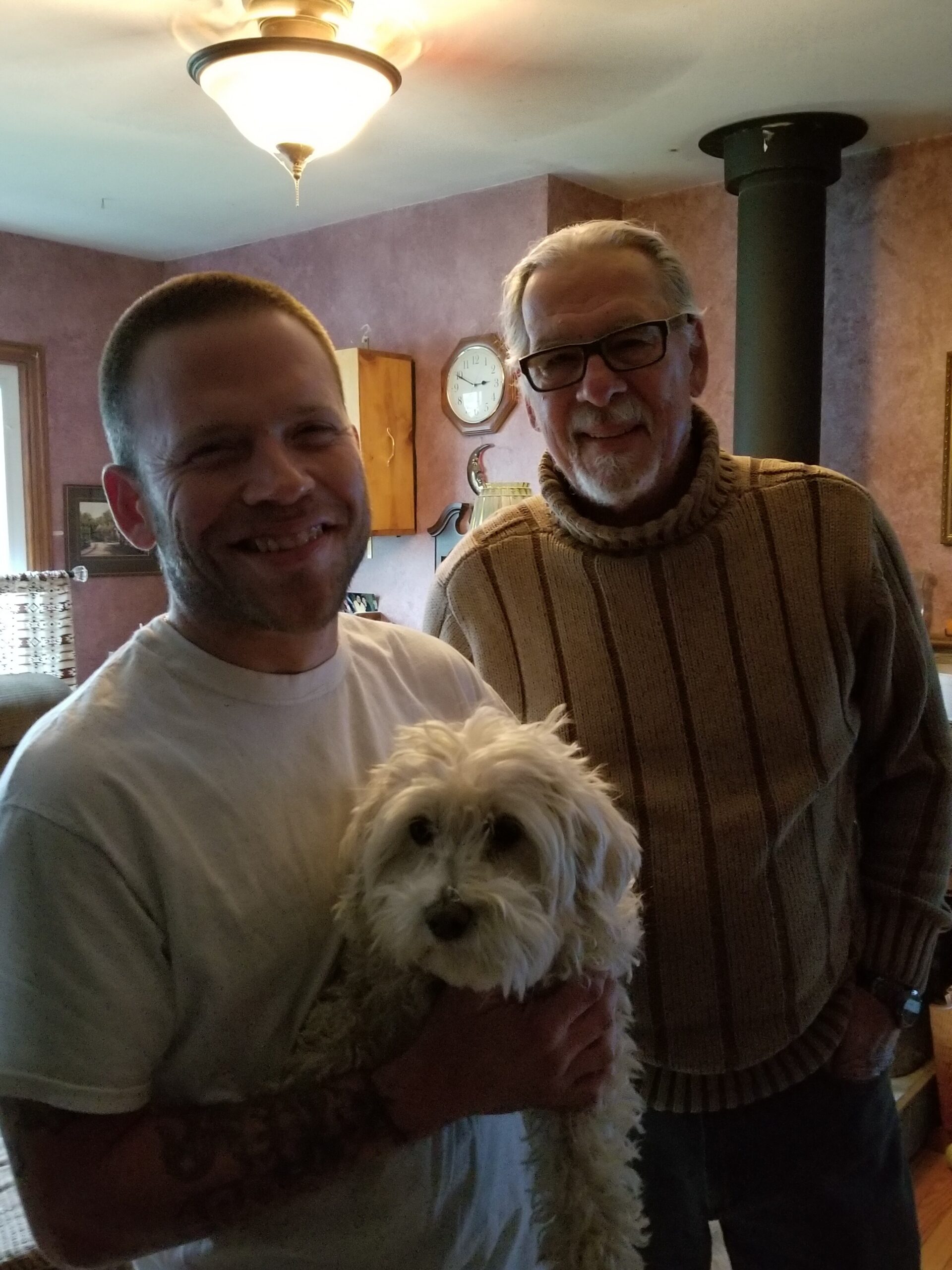 Two men indoors posing with a small fluffy white dog; younger man in a white shirt and older man with glasses and a brown sweater nearby.