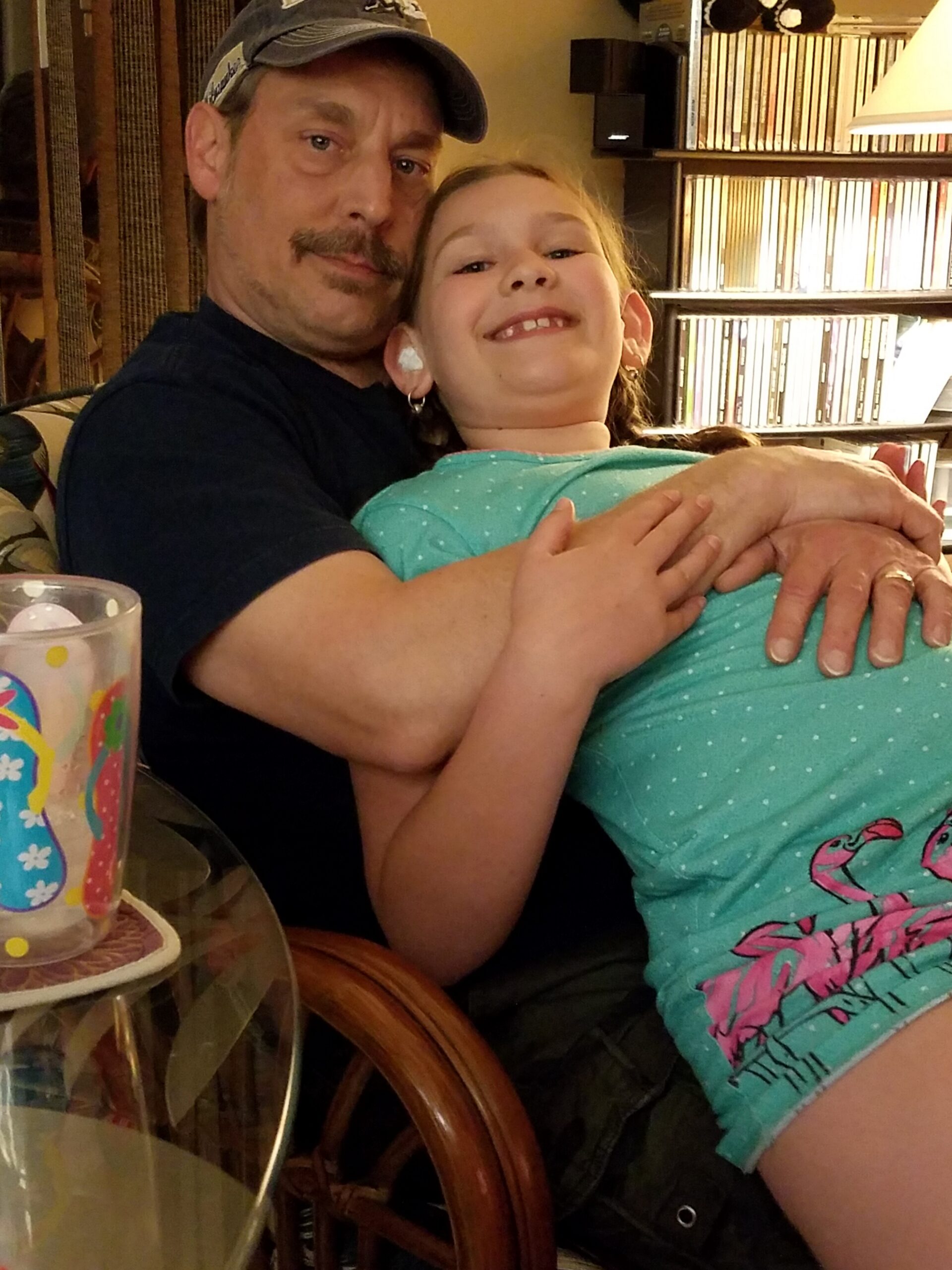 A man wearing a cap holding a smiling young girl in a hug on a living room couch, with bookshelves in the background and a colorful cup on the table nearby.