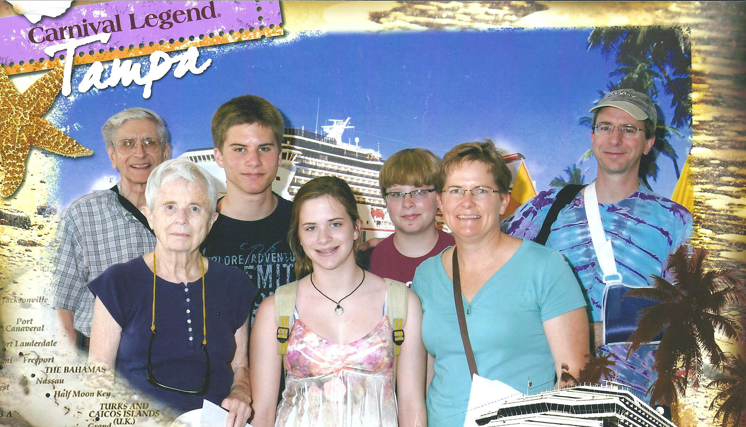 Seven people posing for a group photo in front of a Carnival cruise backdrop featuring a large ship, palm trees, and a starfish.