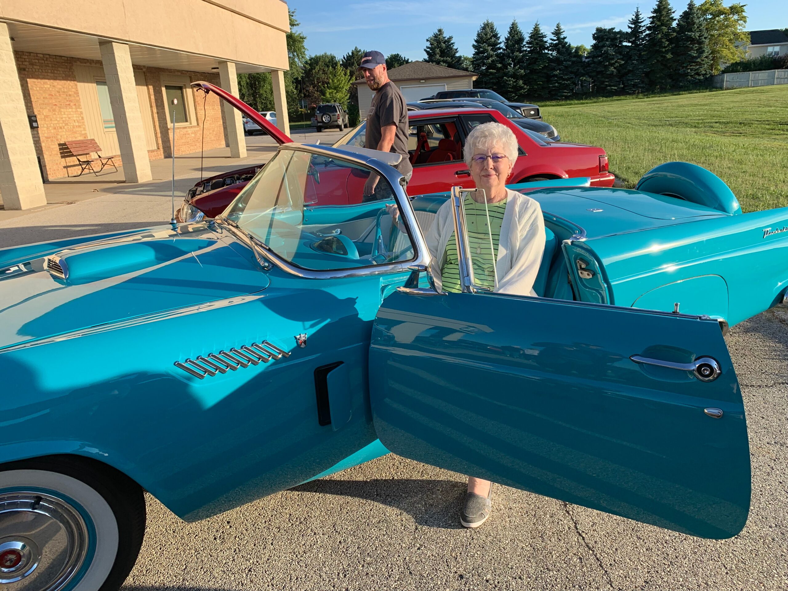 Older woman with white hair and glasses sitting in a turquoise vintage convertible with the door open.
