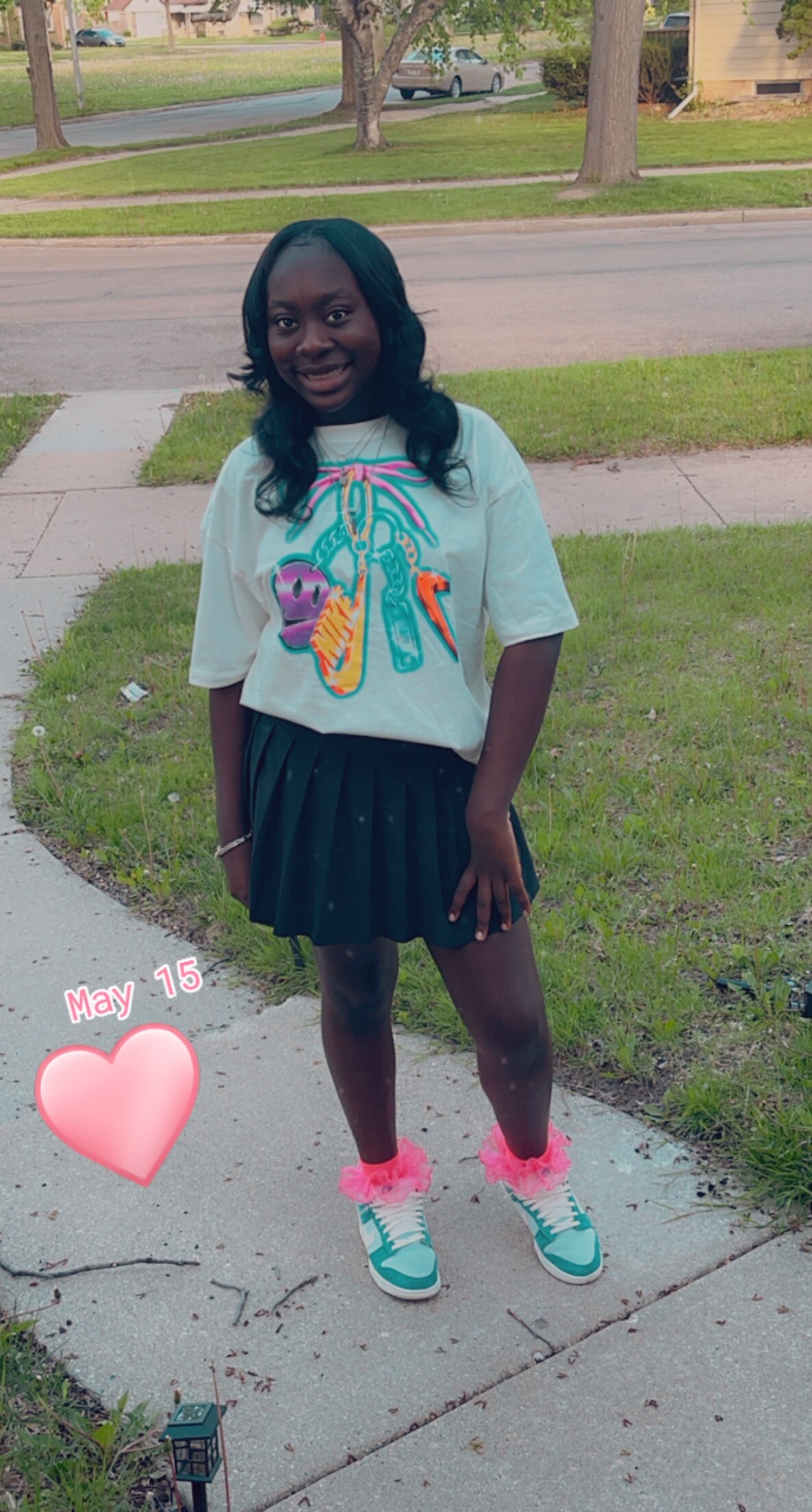 Smiling girl standing on a sidewalk in a suburban street, wearing a colorful graphic T-shirt, black pleated skirt, teal sneakers with pink fluffy socks.