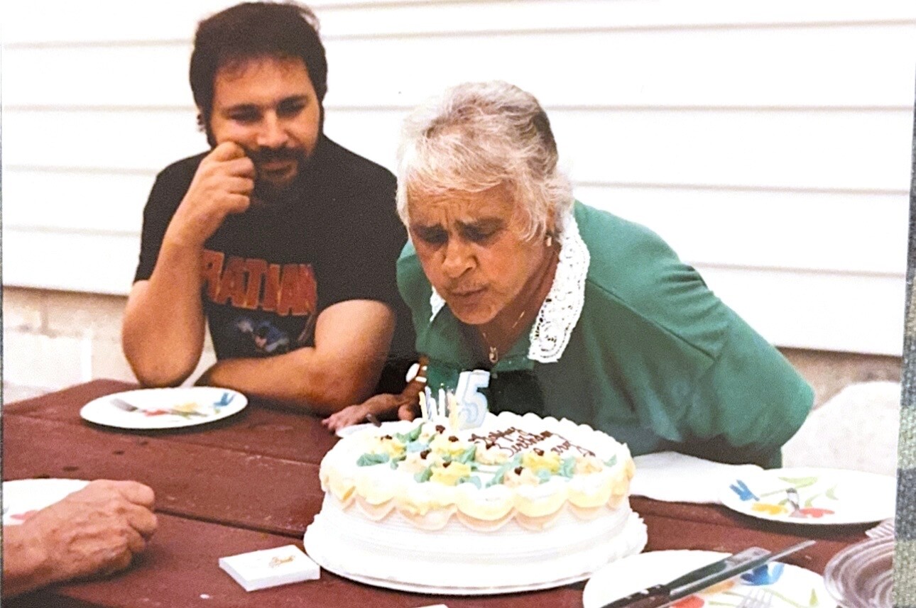 Older woman in a green top leans toward a frosted birthday cake with candles while a man sits nearby at a wooden table outside.