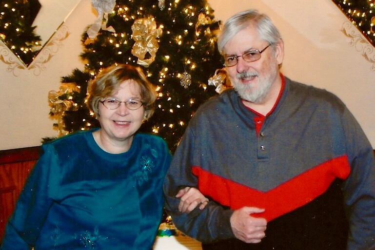 Smiling older couple stands arm in arm in front of a decorated Christmas tree.