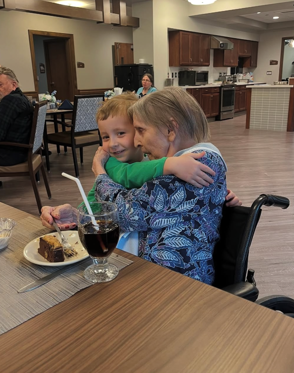 An elderly woman in a patterned blue top embraces a smiling boy at a dining table in a communal kitchen/dining area; cake and a cola are nearby.
