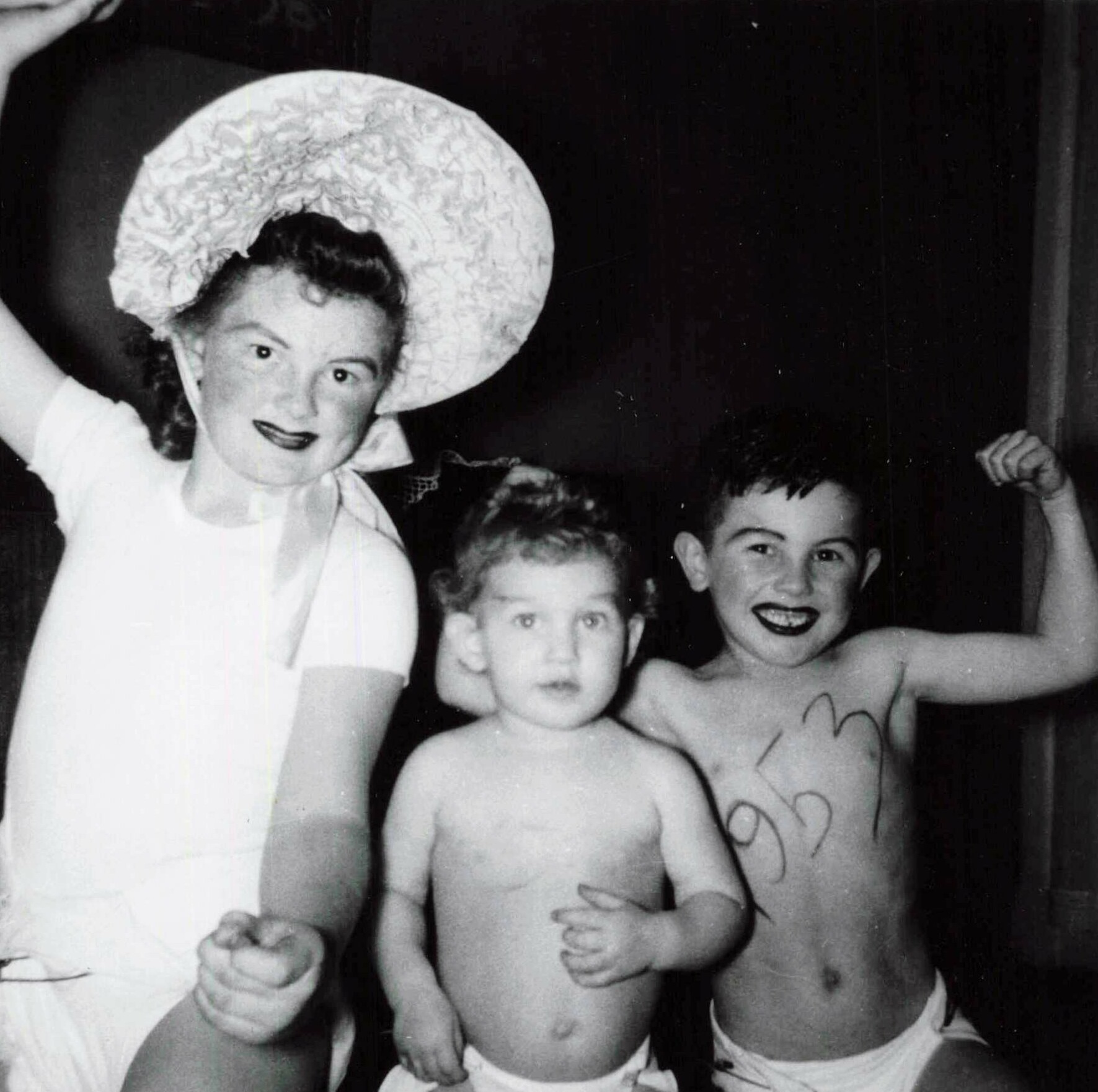 Black-and-white photo of a smiling woman wearing a large patterned hat posing with three shirtless children at a festive event.