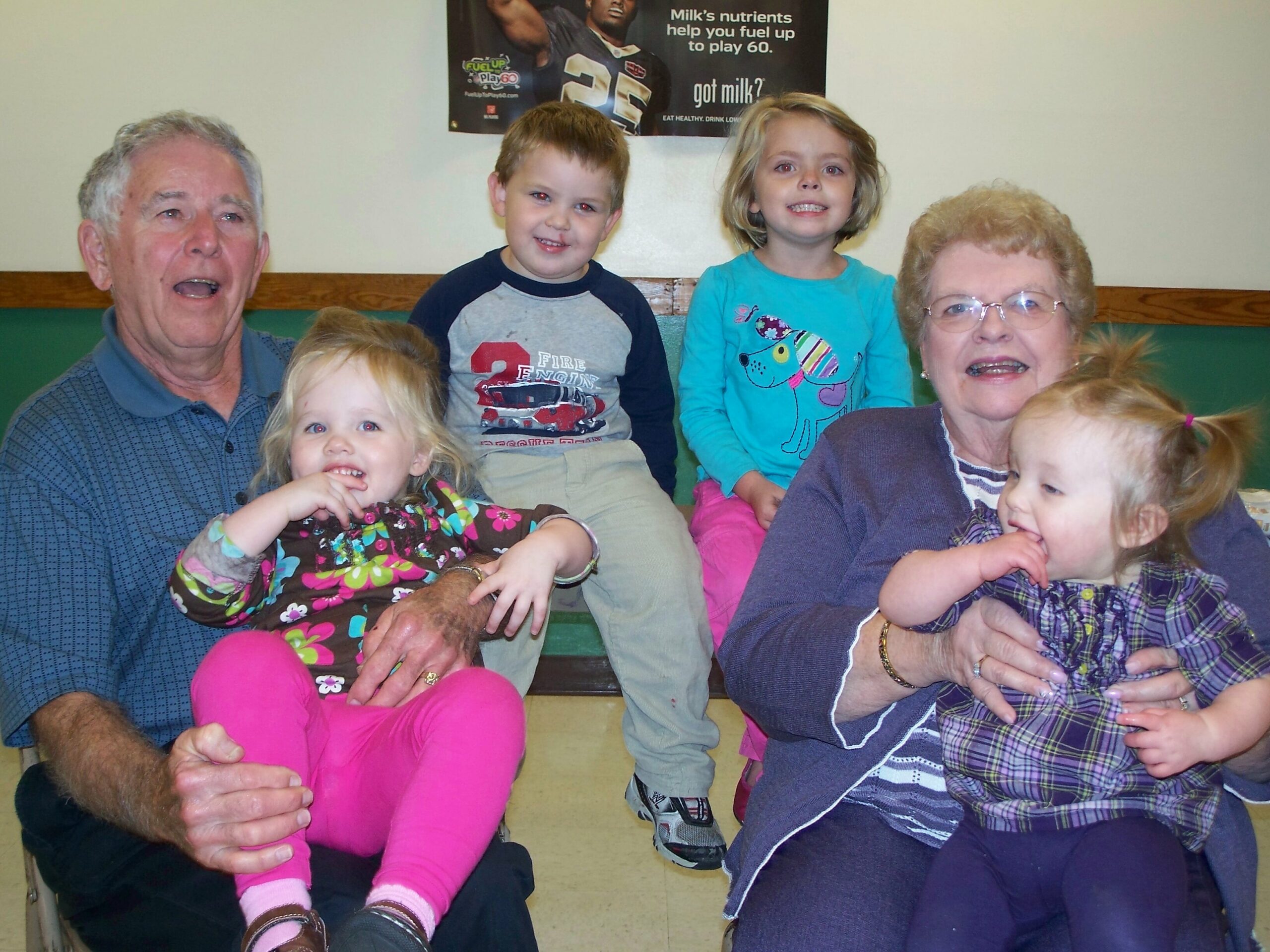 Group of five people posing together indoors: an older man on the left, three children in the middle, and an older woman on the right, all smiling.