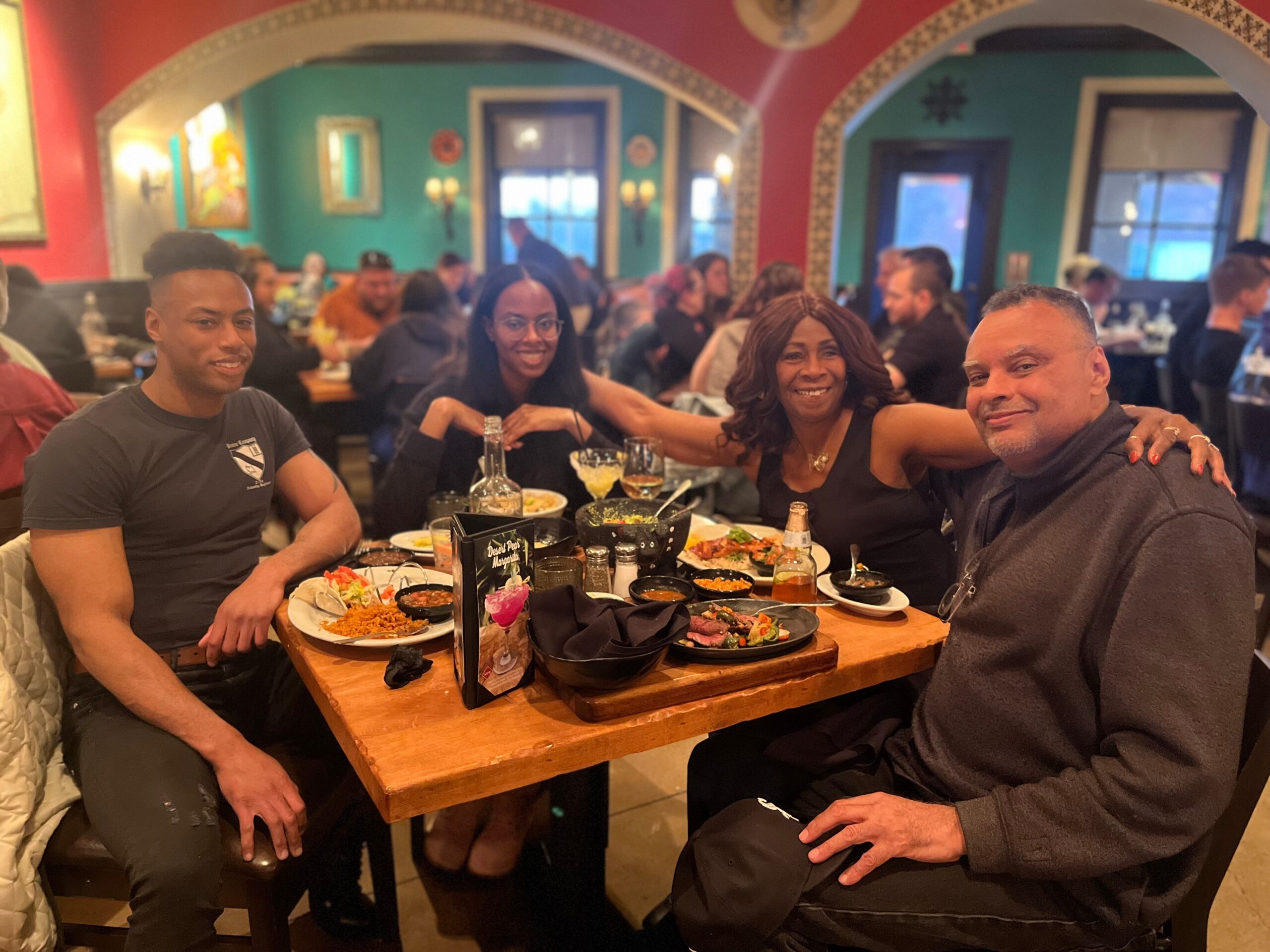 Five adults sit around a wooden table at a colorful restaurant, smiling with plates of food and drinks in front of them.