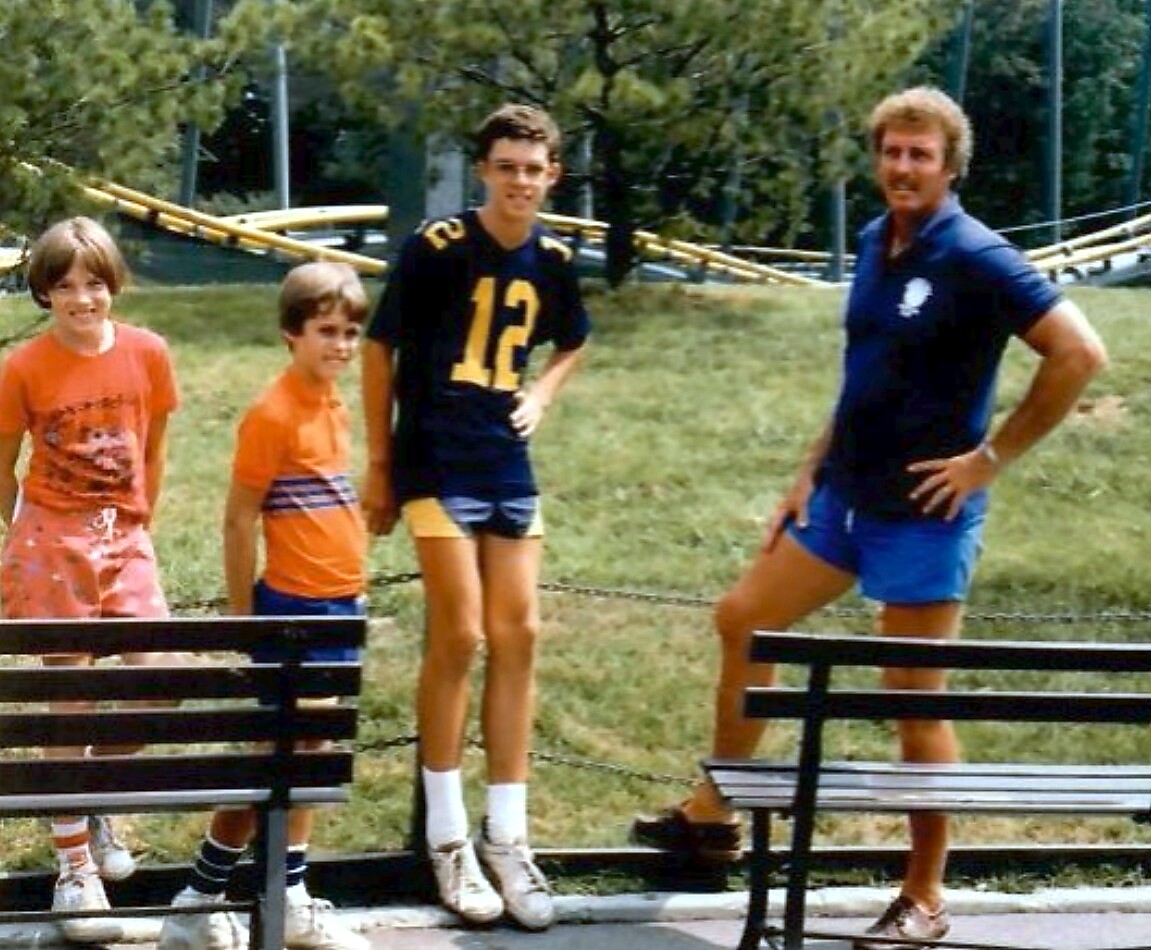 A man in a blue polo shirt and shorts stands with three boys in a park, near benches and a chain fence. They wear casual athletic clothes and sneakers, suggesting a sports practice or game.