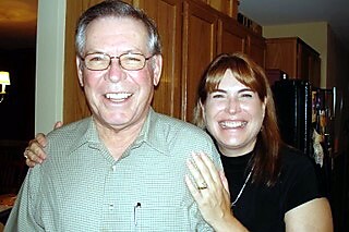 Smiling older man wearing glasses and a younger woman in a black shirt pose together in a home kitchen, arms around each other.