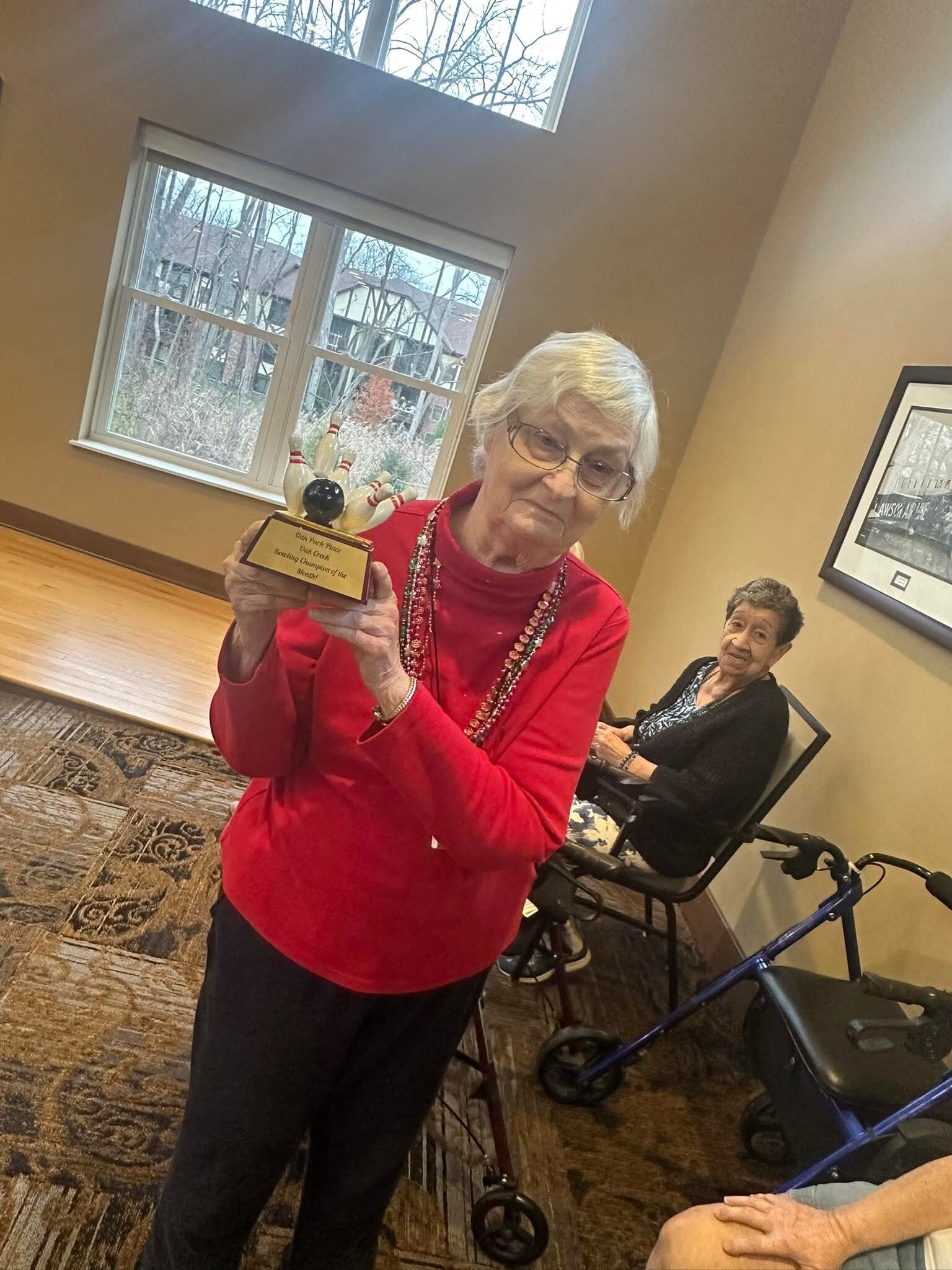 Senior woman in a red top holds a bowling trophy with pins, smiling, as another woman sits in a wheelchair nearby in a lounge area.