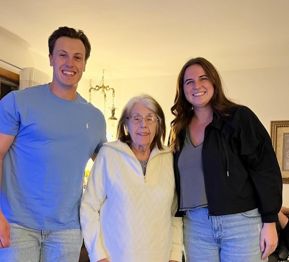 Three generations posing together indoors: a smiling young man in a blue T-shirt, an elderly woman in a cream sweater, and a smiling young woman in a black jacket.