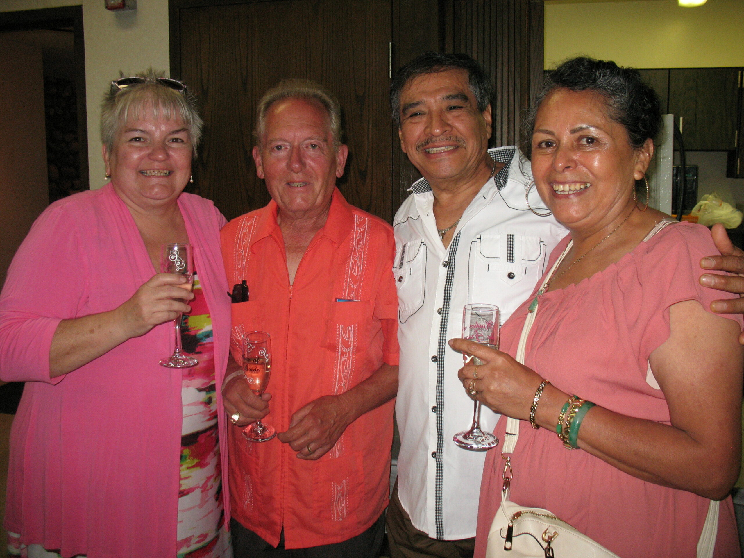 Four adults posing at a party, two women in pink, one man in orange, and another woman in pink, all smiling with drinks.