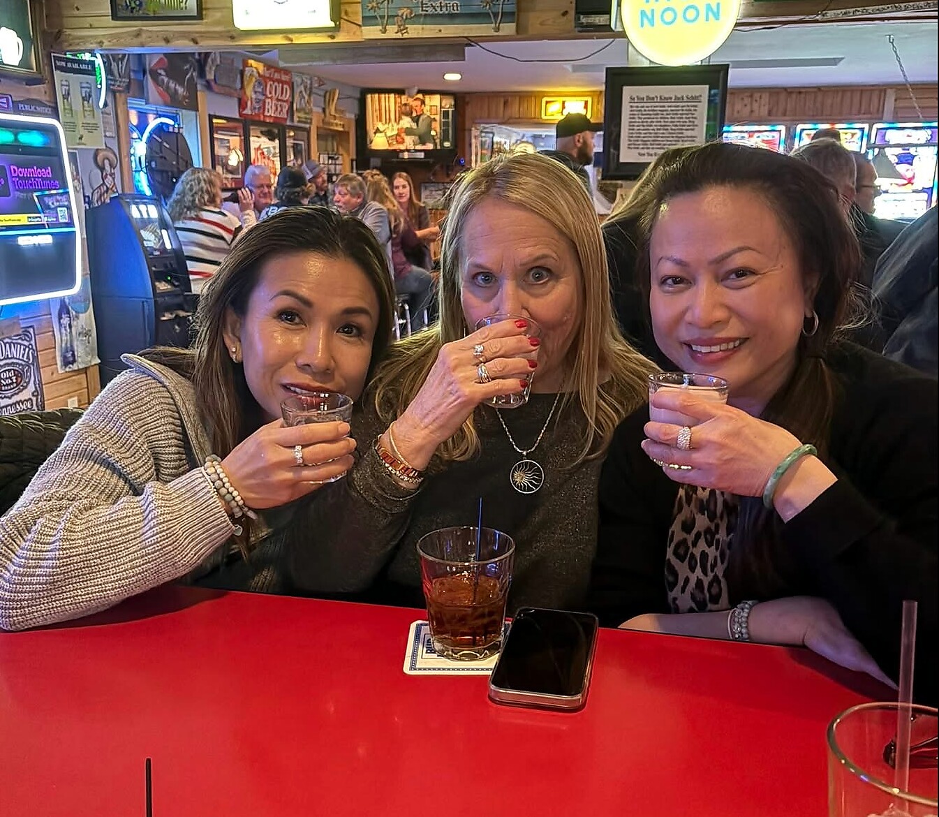 Three women seated at a red-bar counter, raising glasses for a toast in a busy casual bar setting.