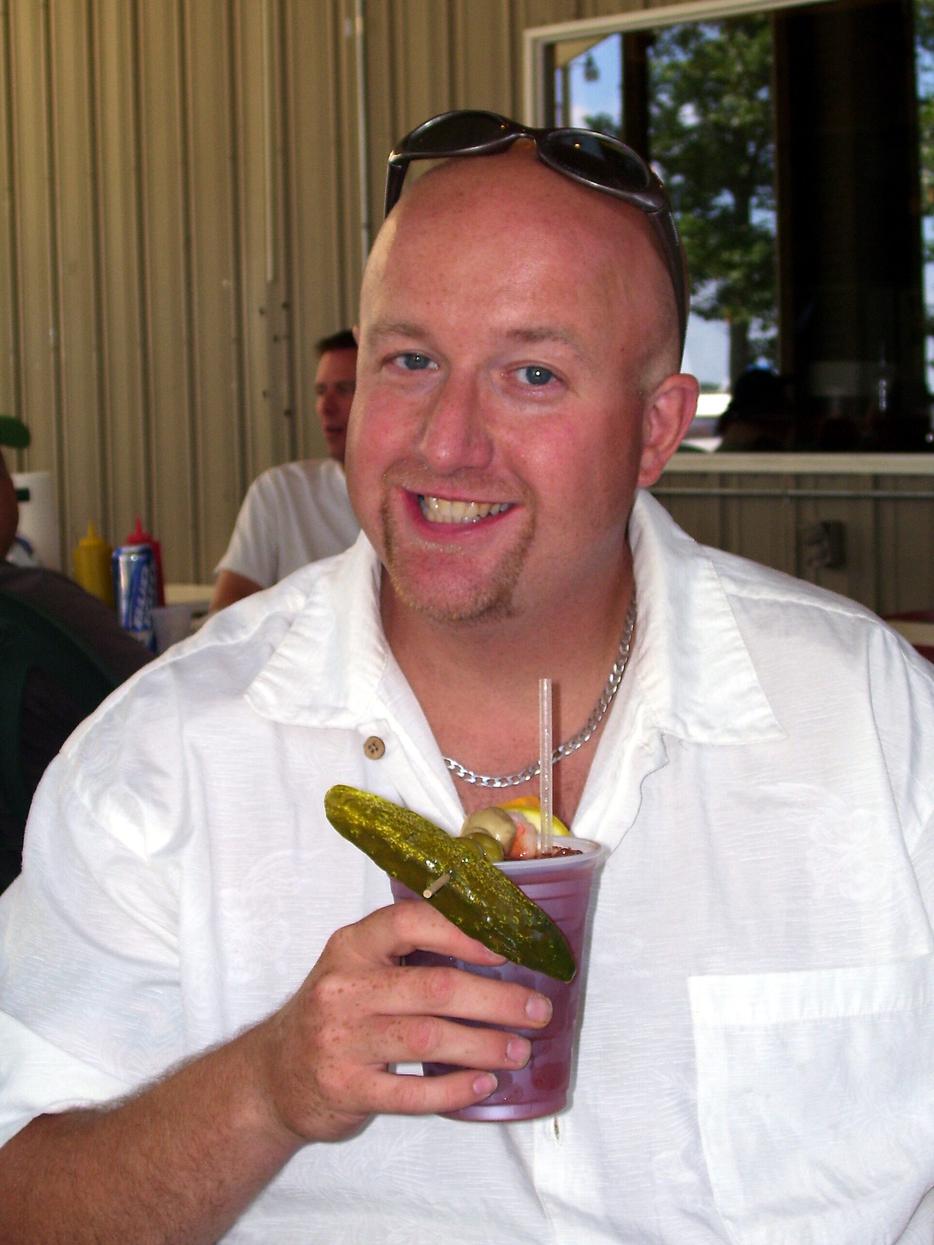 Smiling man in a white shirt holds a purple cocktail with a pickle spear garnish in front of him.