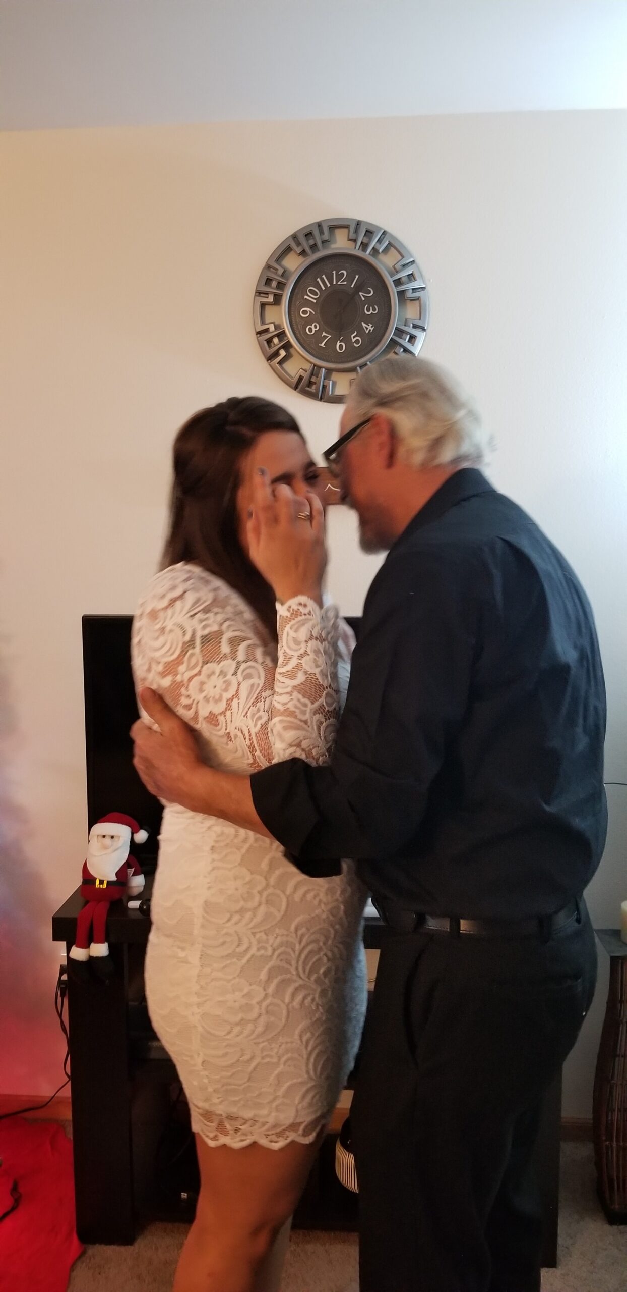 A couple embraces indoors near a wall clock, the woman in a white lace dress and the man in dark clothing.
