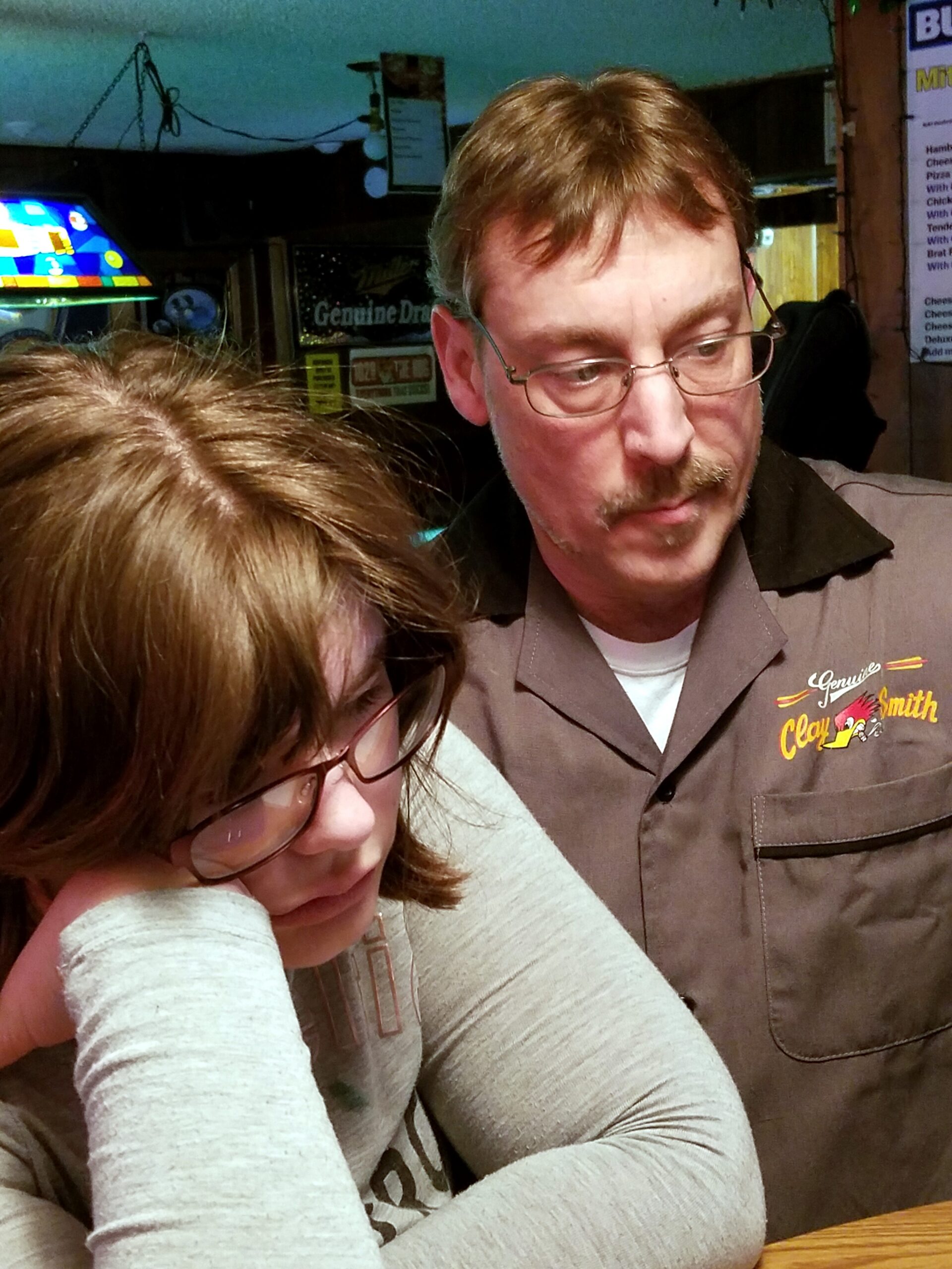 Two people sit closely at a wooden table in a casual restaurant; the man wears glasses and a brown shirt with a logo, looking down as the woman in front rests her head on her arms.