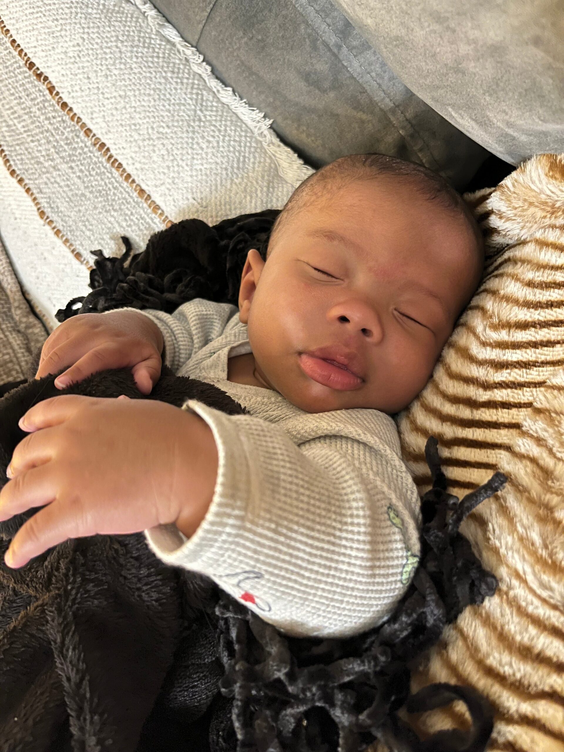 Infant sleeping peacefully while nestled in soft blankets and a striped faux-fur pillow, holding a dark plush toy