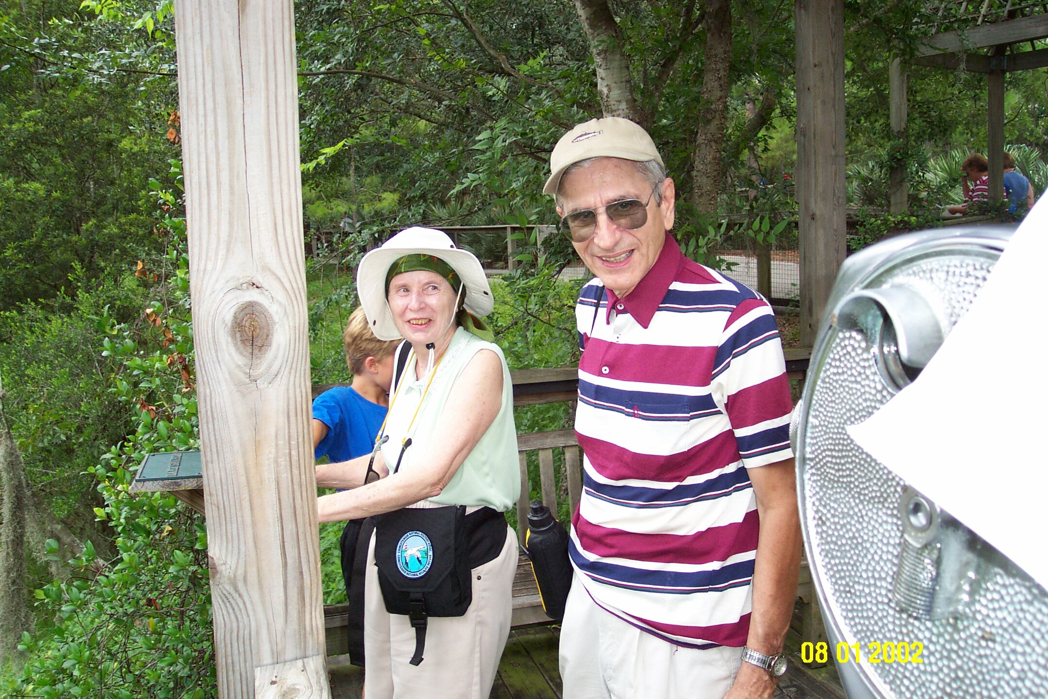 Smiling senior man in a pink, white, and Navy striped polo shirt and beige cap posing beside a woman wearing a white hat, both standing on a wooden deck with greenery around.