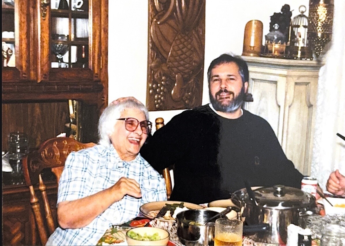 Older woman with white hair and glasses laughing, seated at a dining table with a man beside her during a meal in a cozy room with dishes and drinks nearby.