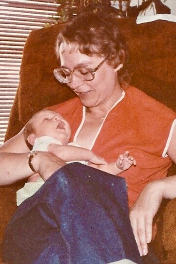 Woman with short hair and glasses cradling a newborn baby in her arms while sitting in a chair, baby wrapped in a light outfit and looking up at her.