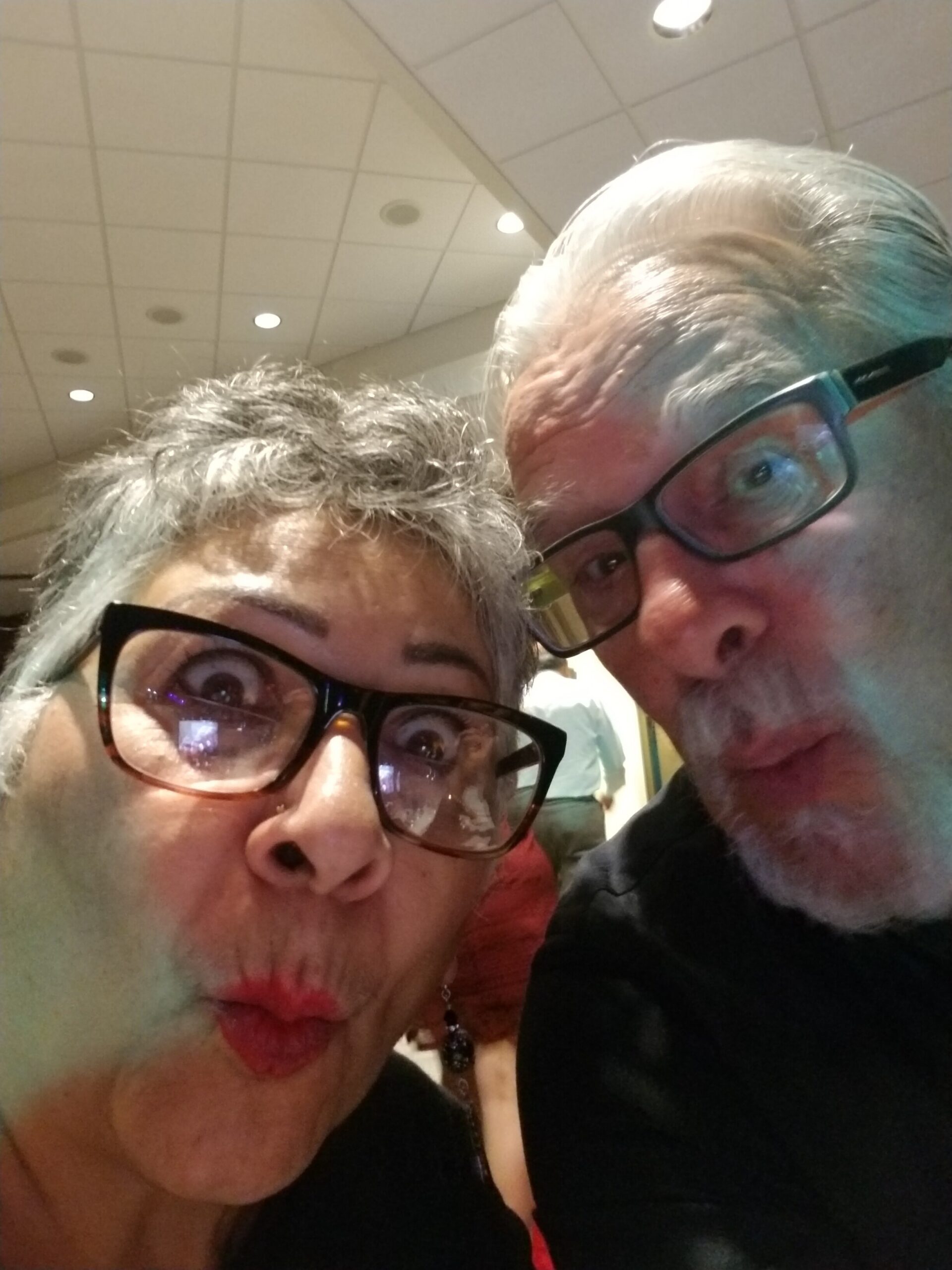 Two people take a playful close-up selfie indoors, both puckering lips at the camera while leaning in together.