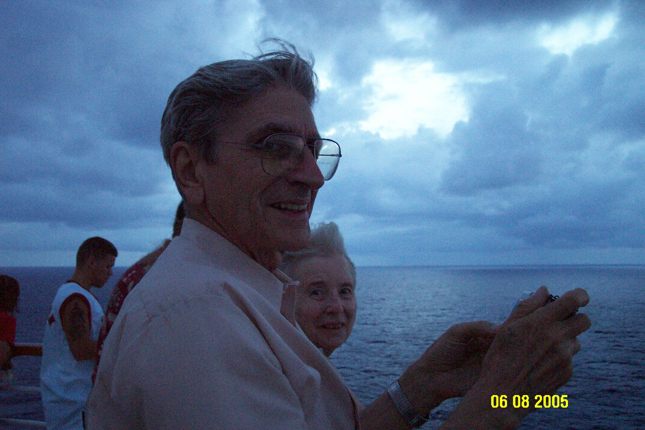 Smiling man wearing glasses on a boat at dusk, with a cloudy sky and sea behind him.