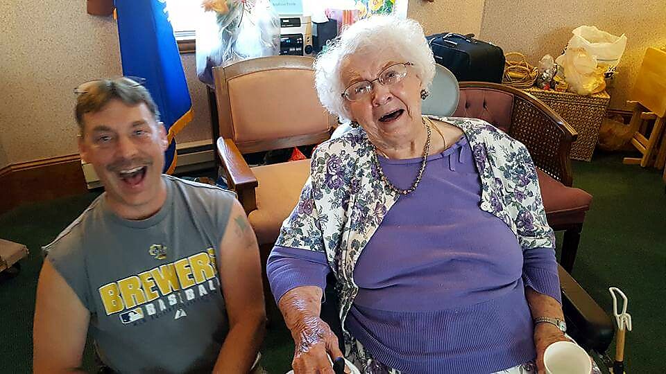 Smiling man in a sleeveless gray Brewers shirt sits next to an elderly woman with white hair and glasses, both laughing in a cozy room with chairs and personal items in the background.