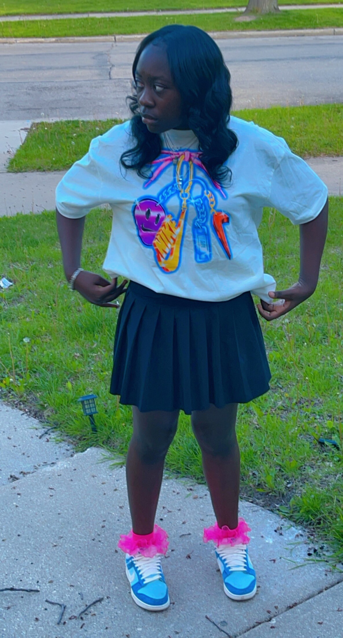 Young person standing on a sidewalk wearing a white graphic T-shirt, black pleated skirt, bright blue sneakers, and pink fluffy socks, looking to the side.
