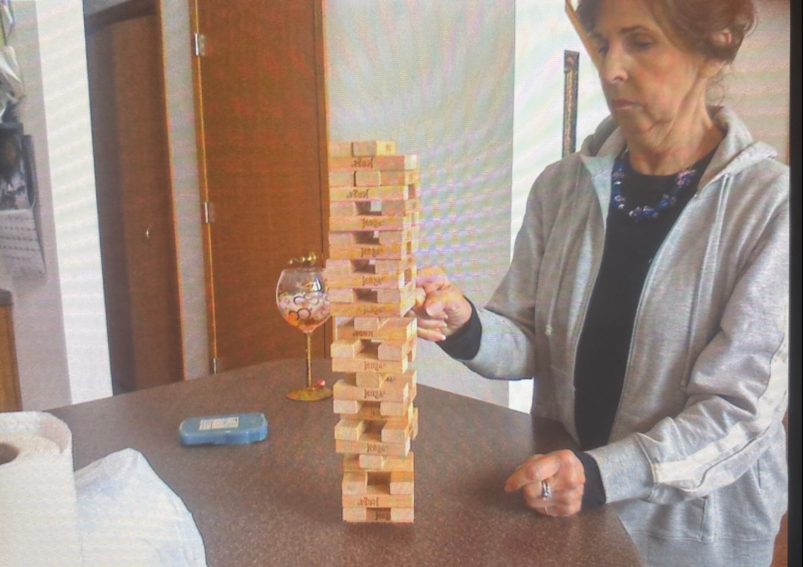 Woman in a gray jacket stacks a Jenga tower on a table, with a decorated glass nearby.